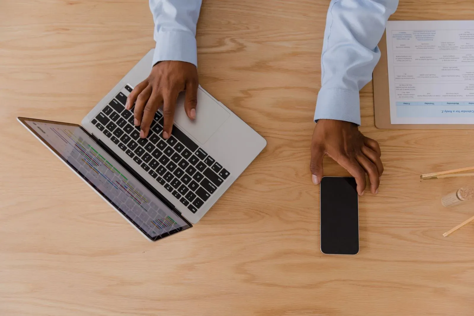 Manos trabajan con un portátil y un teléfono sobre una mesa de madera.